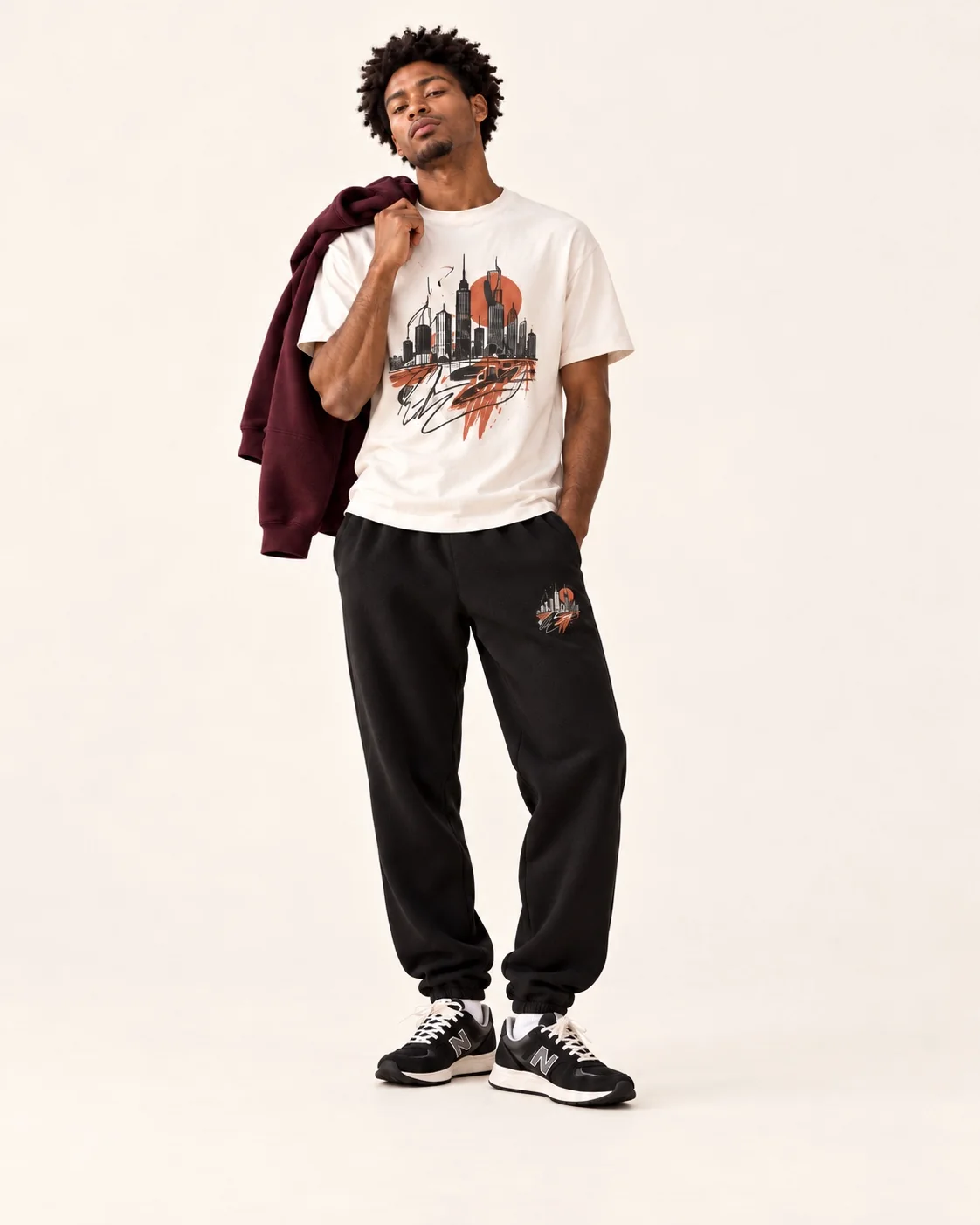 A man in a white graphic tee and black joggers with a small matching print on the leg, a burgundy hoodie slung over his shoulder